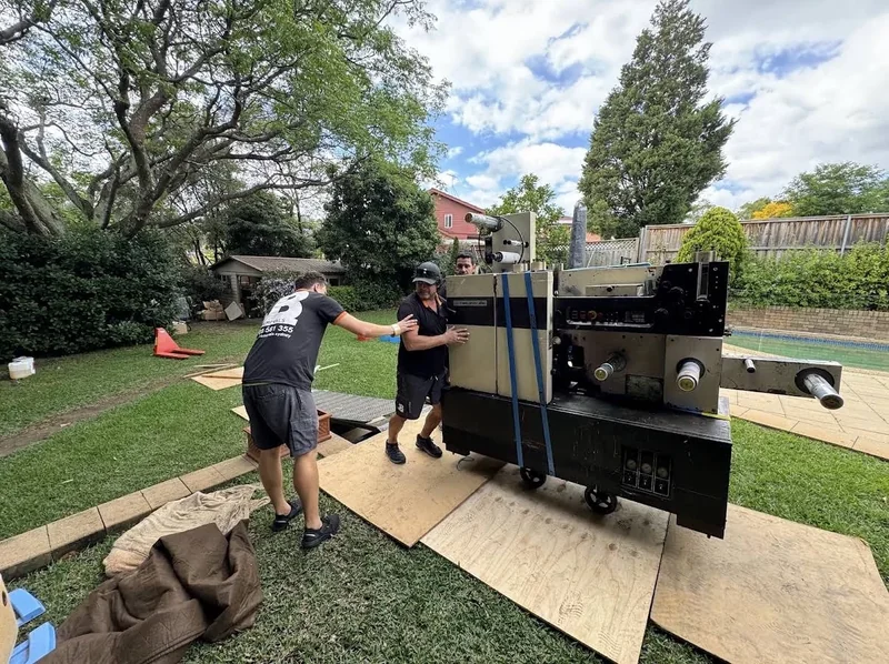 Large sticker-printing machine with blue restraint straps on wheeled supports over sheet material beside a garden and paved area