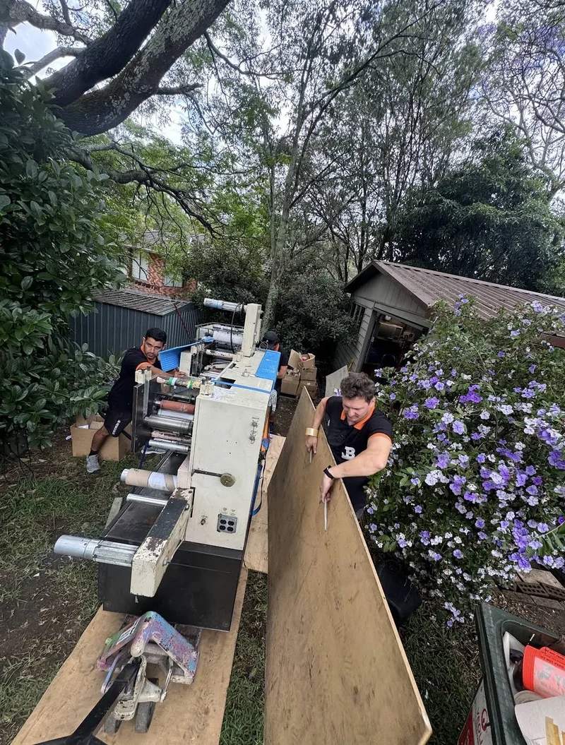JB crew beside a printing machine and a large upright sheet along a confined outdoor route near an outbuilding and garden